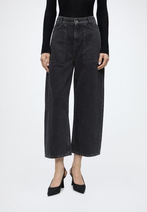 Personne portant un jean noir large et court avec de grandes poches avant, un pull noir côtelé et des escarpins noirs pointus, debout.
