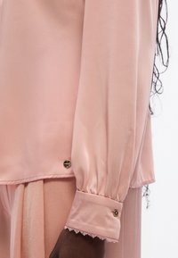 Lyserød bluse lavet af glat stof med en rynket ærme, gyldne knapdetaljer og delikat blondestrimle ved manchetten.