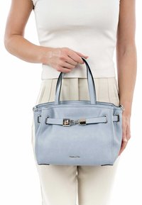 Žena v bílém topu a kalhotách drží světle modrou koženou kabelku se stříbrným uzávěrem před bílým pozadím.