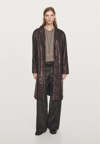 Lange bruine leren jas met een glanzende afwerking, gecombineerd met een grijze top en wijd uitlopende donkere broek. Eenvoudige zwarte knopen en een hangende ketting.