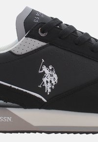 Fekete sportcipő perforált szürke díszítésekkel, fehér póló játékos logóval. Texturált anyag, lapos fűzők, és fehér talp jelzéssel.