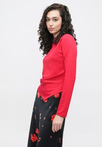 Κόκκινο μακρυμάνικο τοπ με στρογγυλεμένο τελείωμα, συνδυασμένο με μαύρη φούστα με λουλουδάτο μοτίβο. Το σύνολο έχει απαλή υφή.