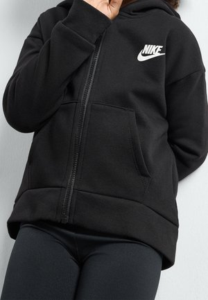 Person iført sort Nike hættetrøje med lynlås og hvidt logo samt sorte leggings står op ad en lys væg, hånd tæt ved ansigtet, overkrop og ben synlige.
