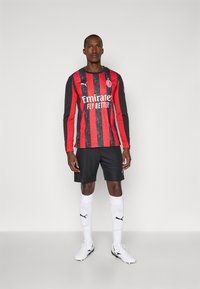 Sort og rød langærmet fodboldtrøje med lodrette striber, prydet med et logo og tekst. Sorte shorts, hvide sokker med logo og matchende fodtøj.