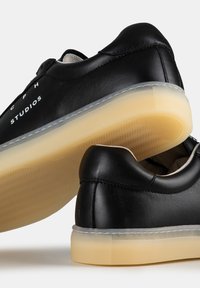 Baskets en cuir noir avec une texture lisse, dotées d'une semelle en caoutchouc translucide et d'une marque blanche. Forme arrondie avec des détails de design minimaux.