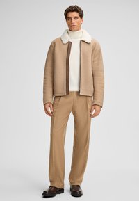 Giacca in shearling color castagna chiaro con colletto in pelliccia sintetica crema, abbinata a un dolcevita bianco a coste e pantaloni marrone chiaro, completata da stivali scuri.