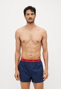 Boxer shorts bleu marine en tissu lisse avec une ceinture élastique rouge portant le logo 'HUGO'. Détail de bouton à l'avant et design minimal.