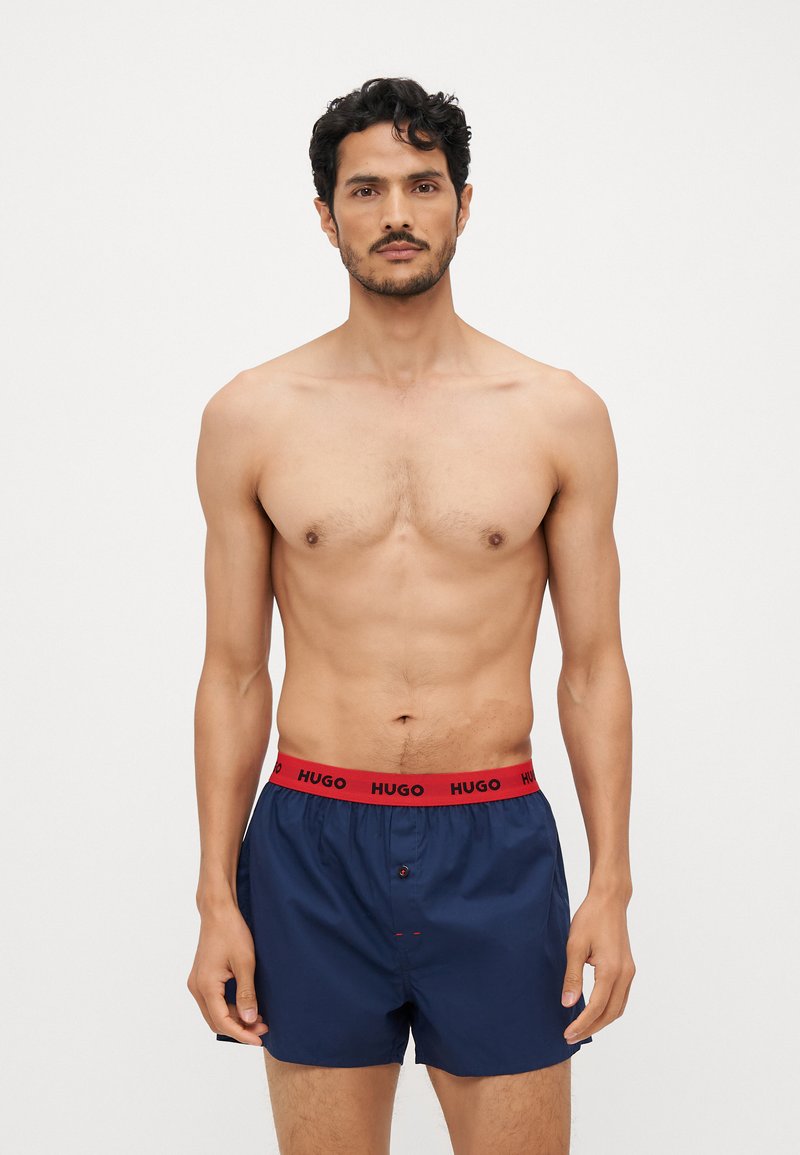 Boxer shorts bleu marine en tissu lisse avec une ceinture élastique rouge portant le logo 'HUGO'. Détail de bouton à l'avant et design minimal.