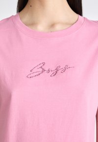 Pink cotton t-shirt with a round neckline featuring sparkling embellishment in cursive script across the chest. Smooth texture.