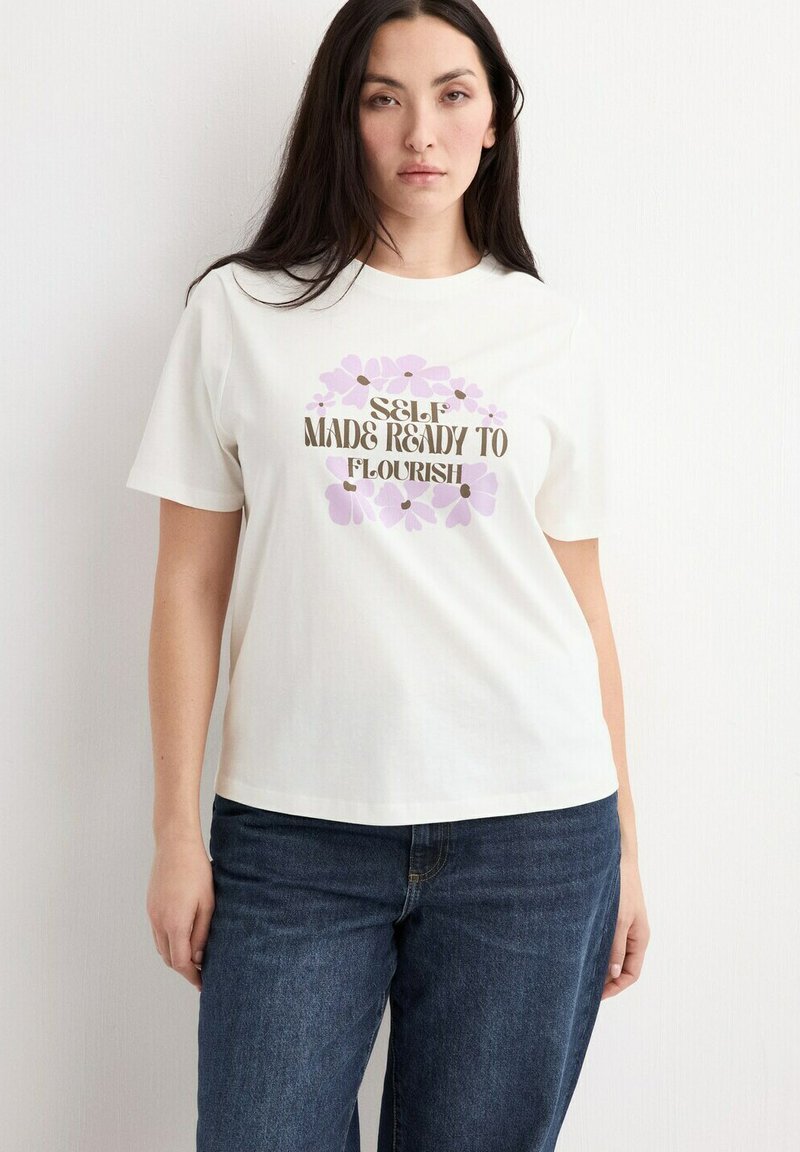 Femme portant un t-shirt blanc avec des fleurs violettes et le texte "Self Made Ready to Flourish", associée à un jean bleu, debout contre un mur blanc.