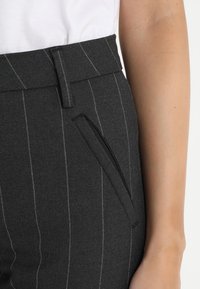 Detailní záběr osoby nosící tmavě šedé kalhoty s proužky, boční kapsou a poutky na pásek, v kombinaci s bílým topem.