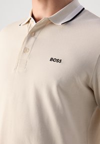 Beige poloskjorte med svart kantet hvit krage, tre beige knapper, og "BOSS" logo brodert i svart på venstre bryst.