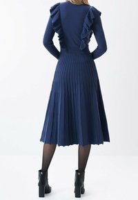 Abito a maglia a coste di colore blu navy con pieghette verticali e dettagli di rouches lungo le spalle, che termina a metà polpaccio. Stivali alla caviglia neri con tacco.