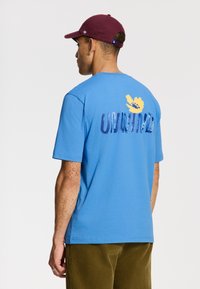 Camiseta de algodón azul de manga corta con la palabra "UNWIND" en texto morado en negrita y un gráfico de una flor amarilla en la parte posterior. Pantalones de pana olivo visibles.
