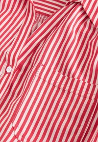 Gros plan d'une chemise rayée verticalement rouge et blanche montrant le col, la poche et un bouton blanc sur un tissu léger.