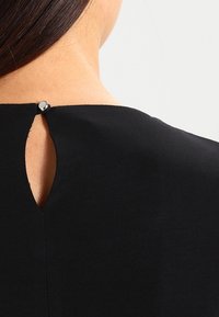 Vêtement noir avec une ouverture en trou de serrure dans le dos, sécurisé par un bouton pression en métal. Texture lisse, design simple.