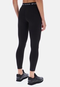 Czarne legginsy sportowe o gładkiej fakturze, z wysokim stanem i białym logo marki, noszone z czarnymi butami sportowymi.