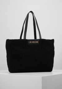 Svart tote-väska gjord av texturerad fleece, med släta svarta läderremmar och en märkesplakett på framsidan. Enkel rektangulär form.