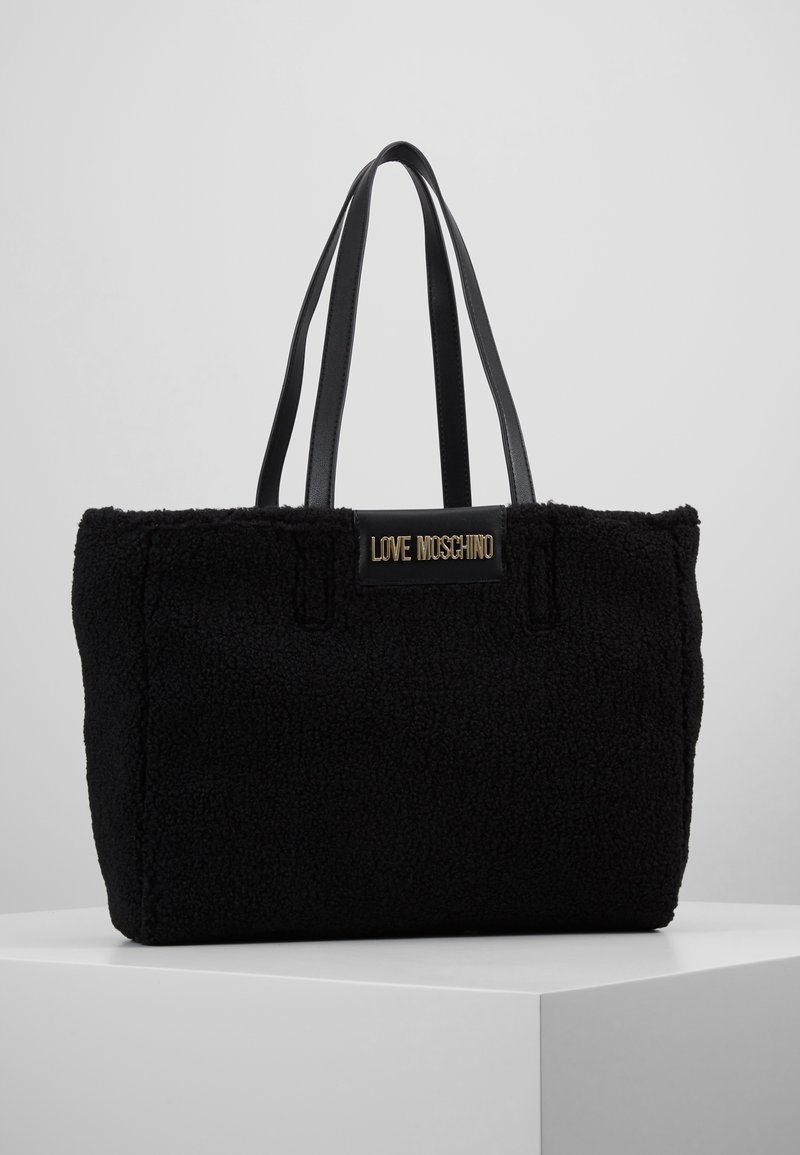 Svart tote-väska gjord av texturerad fleece, med släta svarta läderremmar och en märkesplakett på framsidan. Enkel rektangulär form.