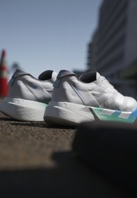Een paar witte sportschoenen met blauw en groene accenten, geplaatst op asfalt onder een heldere hemel naast een wazige rode bouwkegel en een gebouw.