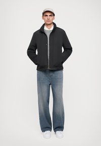 Svart jacka med hög krage, bärs över en grå tröja. Ljusblå jeans, vita sneakers och en beige keps fulländar outfiten.