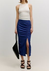 Weißes Trägershirt mit körperbetontem Design, kombiniert mit einem blauen, gerafften Midirock mit einem seitlichen Schlitz und strukturiertem Stoff. Schwarze Crossover-Tasche und Sandalen.
