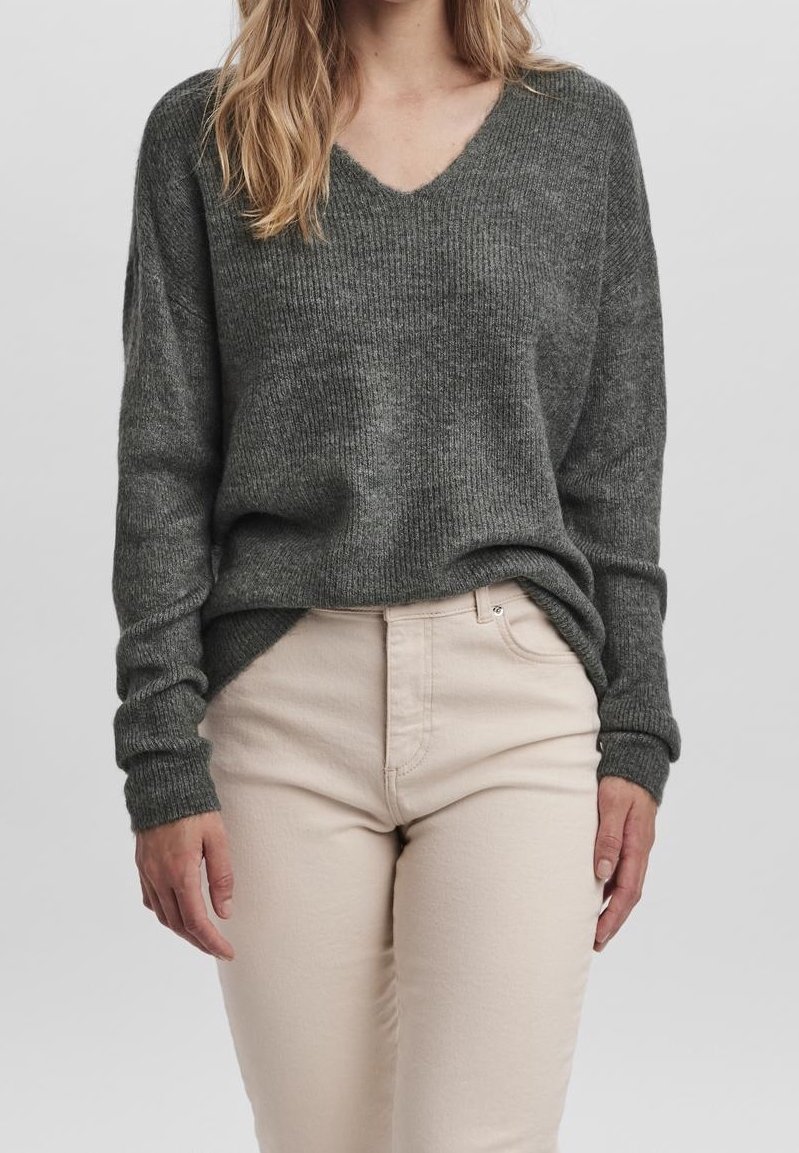 Femme portant un pull à col en V côtelé gris foncé, légèrement rentré dans un pantalon beige clair, sur un fond uni.