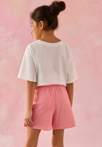 T-shirt cropped en coton blanc et shorts en vichy rose avec une taille élastique. Personne de dos sur un fond rose doux.
