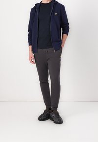 Sudadera de cremallera azul marino con cordones, camiseta oscura, joggers de carbón con un logo y zapatillas deportivas negras con detalles en marrón.