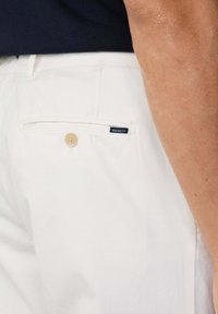Pantaloni di cotone bianchi con una tasca posteriore, dotati di chiusura a bottone e etichetta blu "HACKETT". Il design è semplice e sartoriale.