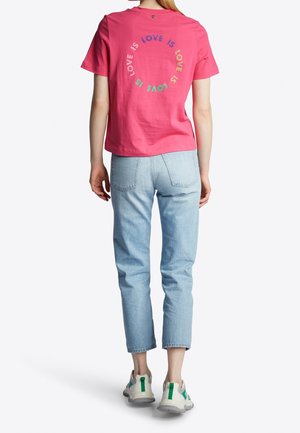 Kvinde iført en knaldpink T-shirt med flerfarvet "LOVE IS" cirkeltekst, lyseblå korte jeans og hvide sneakers med grønne detaljer.