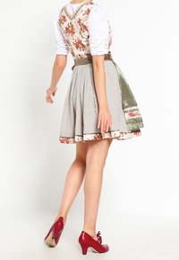 Kobieta w sukience dirndl w kwiatowy wzór z kraciastą spódnicą i oliwkowym fartuszkiem, w połączeniu z czerwonymi butami na obcasie, stojąca na białym tle.