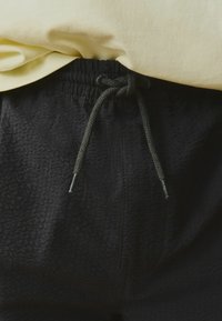 Jogging noir texturé avec une taille à cordon de serrage, présentant un cordon tressé vert et des embouts métalliques. Une chemise jaune pâle est partiellement visible au-dessus.