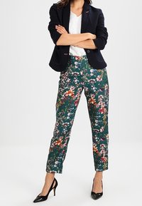 Floral trousers in dark green fabric, featuring various colors and patterns of flowers. Paired with a black blazer and high-heeled shoes.