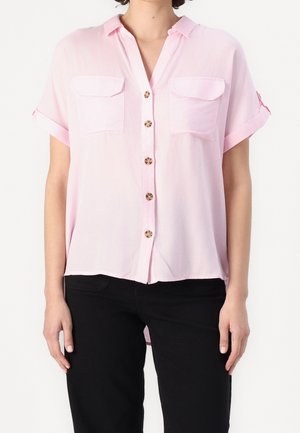 Kvinde iført en lyserød kortærmet skjorte med to brystlommer og sorte bukser, stående mod en hvid baggrund.