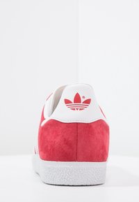 Červené semišové tenisky s bílými akcenty. Mají plochou, texturovanou podešev a logo na zadní straně. Klasický nízký design, vhodné pro volný čas.