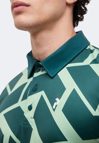 Άνδρας που φοράει πράσινο πόλο με λαιμόκοψη σκούρου πράσινου και ανοιχτό πράσινο γεωμετρικό μοτίβο, κουμπωμένο στην κορυφή.