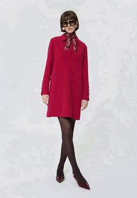 Kobieta w czerwonej sukience z długim rękawem i zapięciem na guziki, czarnych rajstopach, burgundowych szpilkach, wełnianym szalu w kratę i dużych czarnych okularach przeciwsłonecznych.