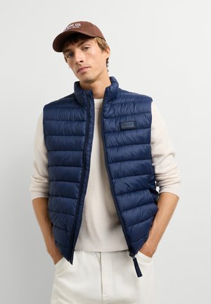 Un homme pose avec un gilet matelassé bleu marine sur un pull crème, un pantalon blanc et une casquette de baseball marron, les mains dans les poches.