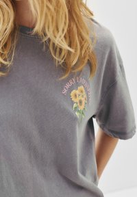 Šedé bavlněné tričko s květinovým vzorem se slunečnicemi a růžovým nápisem "OMLOUVÁM SE, ŽE JSEM TO POKAZIL." Krátké rukávy a volný střih.