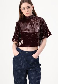 Burgundski crop top sa šljokicama, visokog ovratnika i kratkim rukavima, u kombinaciji s tamnim trapericama koje imaju srebrne ukrase i bočne džepove.