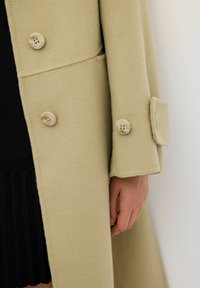 Cappotto di lana khaki con una texture liscia, caratterizzato da bottoni beige e un design strutturato. Dettaglio dei bottoni visibile sul polsino.