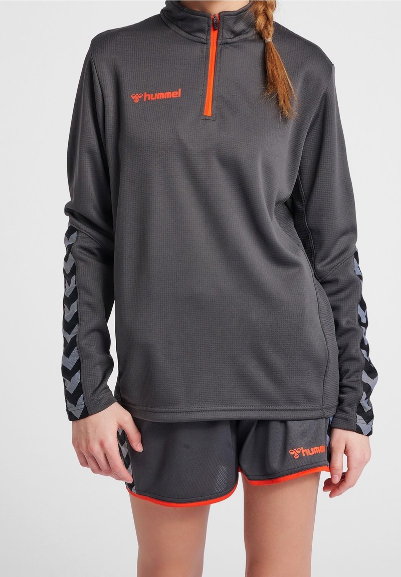 Suéter atlético gris con un alto cuello, cierre de cuarto y cremallera naranja. Presenta un patrón en chevrón de contraste negro y gris en las mangas.