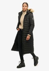 Abrigo puffer negro largo con capucha, que presenta un borde de piel sintética, acolchado horizontal y dos bolsillos laterales. Combinado con un suéter beige de punto trenzado y jeans negros.