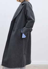 Dark grey wool coat with a notched collar, double buttons, oversized fit, and side pockets. Blue shirt cuffs are visible from underneath.