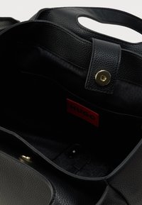 Interiorul unei genți de mână din piele neagră cu granulație, cu un singur buzunar, închidere cu capse aurii și o etichetă roșie de brand vizibilă. Căptușeală textilă netedă.