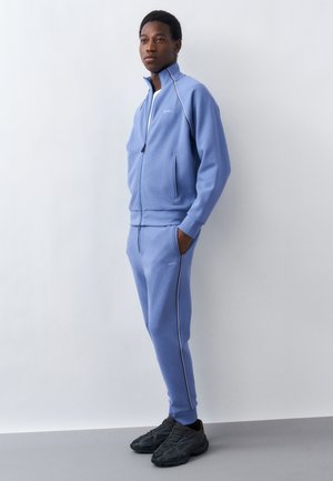 Νέος άνδρας με ανοιχτό μπλε μπουφάν με φερμουάρ και ταιριαστό παντελόνι φόρμας, φορώντας μαύρα αθλητικά παπούτσια, στέκεται δίπλα σε ανοιχτό γκρι τοίχο.