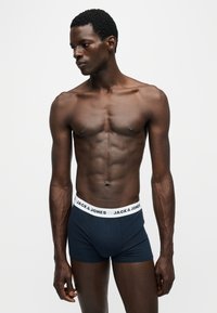 Homme en forme à la peau foncée portant un boxer bleu marine Jack & Jones, debout devant un fond clair uni et regardant sur le côté.
