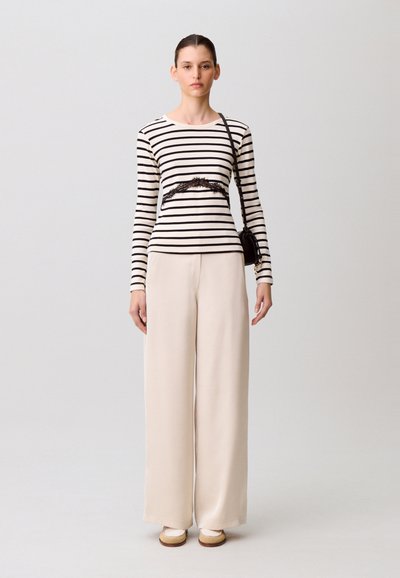 Femme portant un haut à manches longues rayé avec détail en dentelle, un pantalon large beige, des chaussures beiges et un sac à bandoulière noir, posant devant un fond uni.