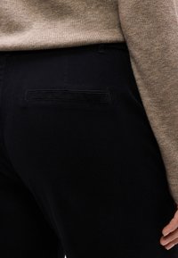 Κοντινό πλάνο ατόμου που φοράει μαύρο παντελόνι με πίσω τσέπη με φάσα και μπεζ πλεκτό πουλόβερ, με το χέρι να ακουμπά κοντά στον γοφό.